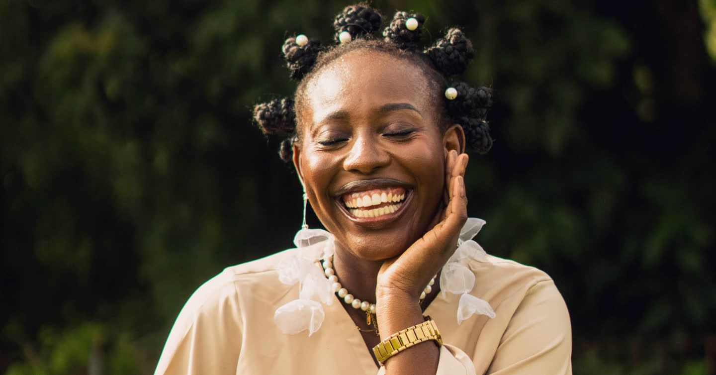 Woman laughing outdoors with eyes closed, hand on cheek, wearing pearl accessories and a beige blouse with clear skin