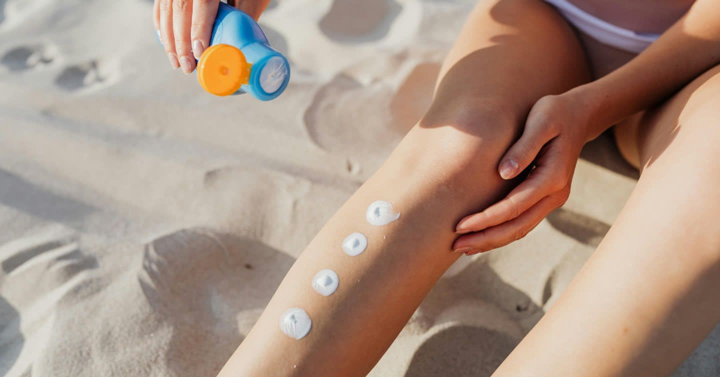 Woman applying sunscreen from a blue bottle onto her leg while sitting on sandy beach