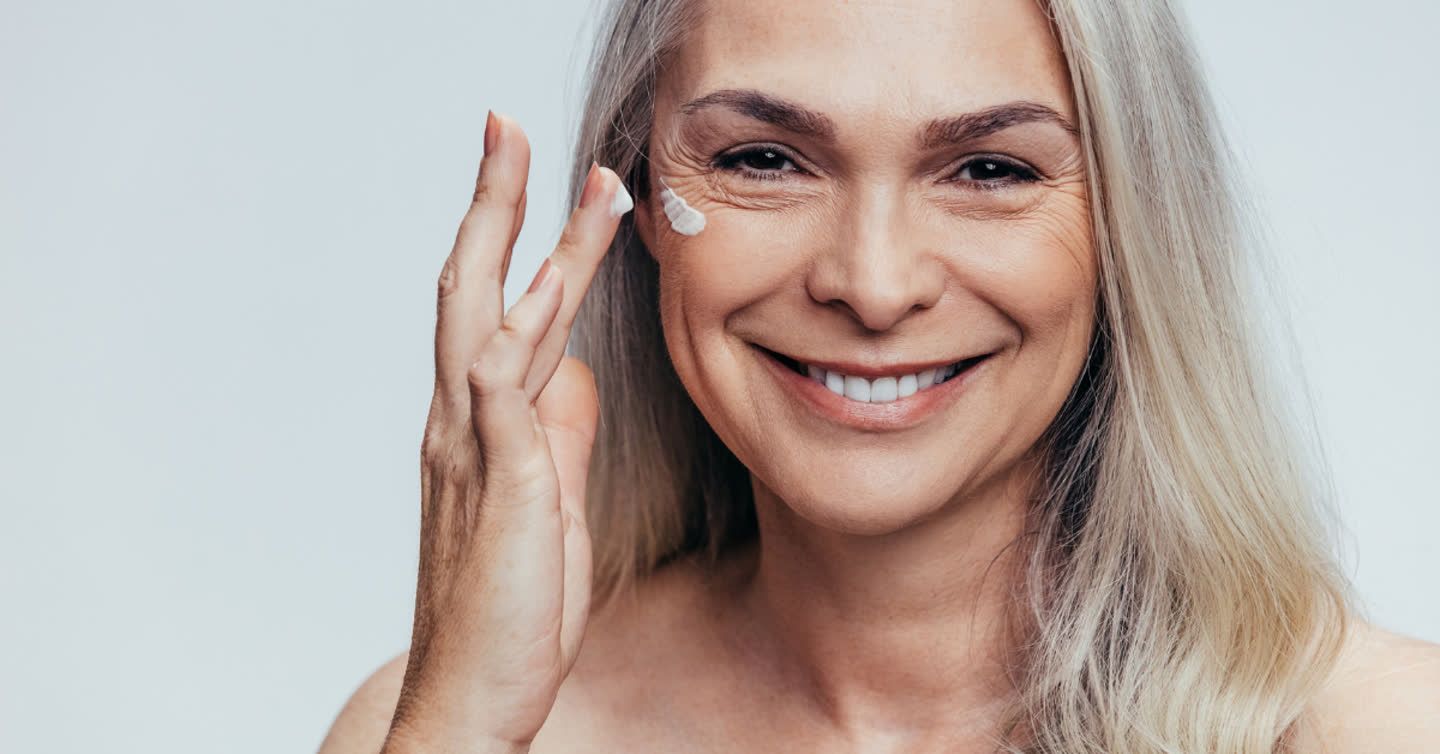 Smiling mature woman applying moisturising cream to her cheek against a light grey background