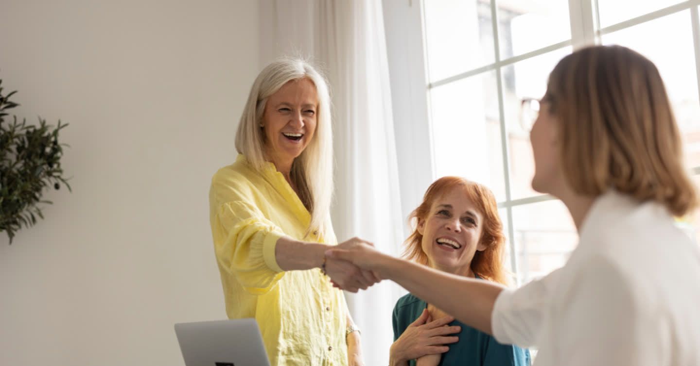 Smiling woman with long grey hair shaking hands with a colleague in a bright office while a red-haired woman looks on