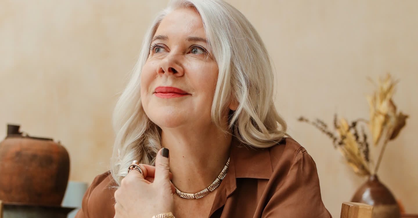 Woman with silver-blonde hair wearing a gold necklace and brown blouse, smiling and gazing upward in a warm-toned room