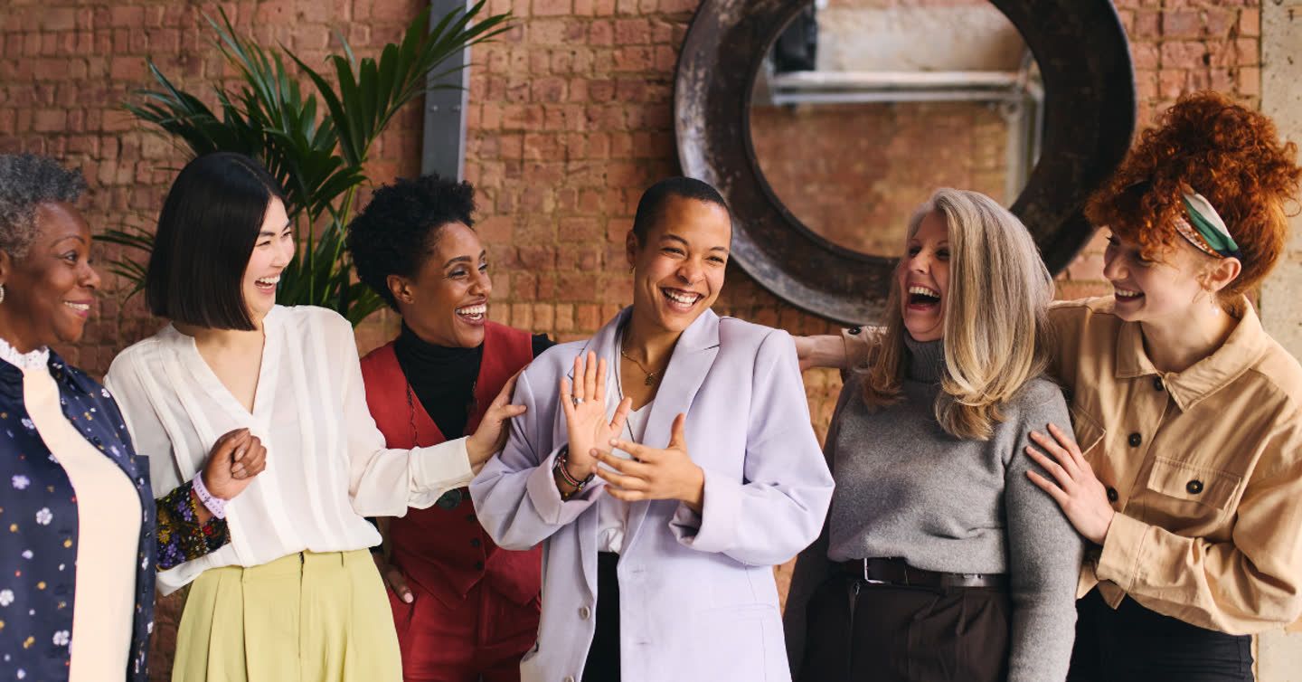 Diverse group of six women laughing together in front of an exposed brick wall with a round mirror