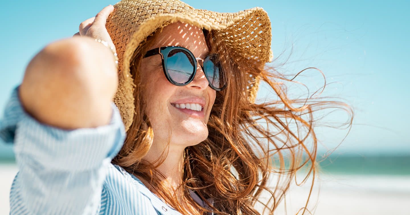 Smiling woman wearing sunglasses and a straw hat on a sunny beach with auburn hair blowing in the wind