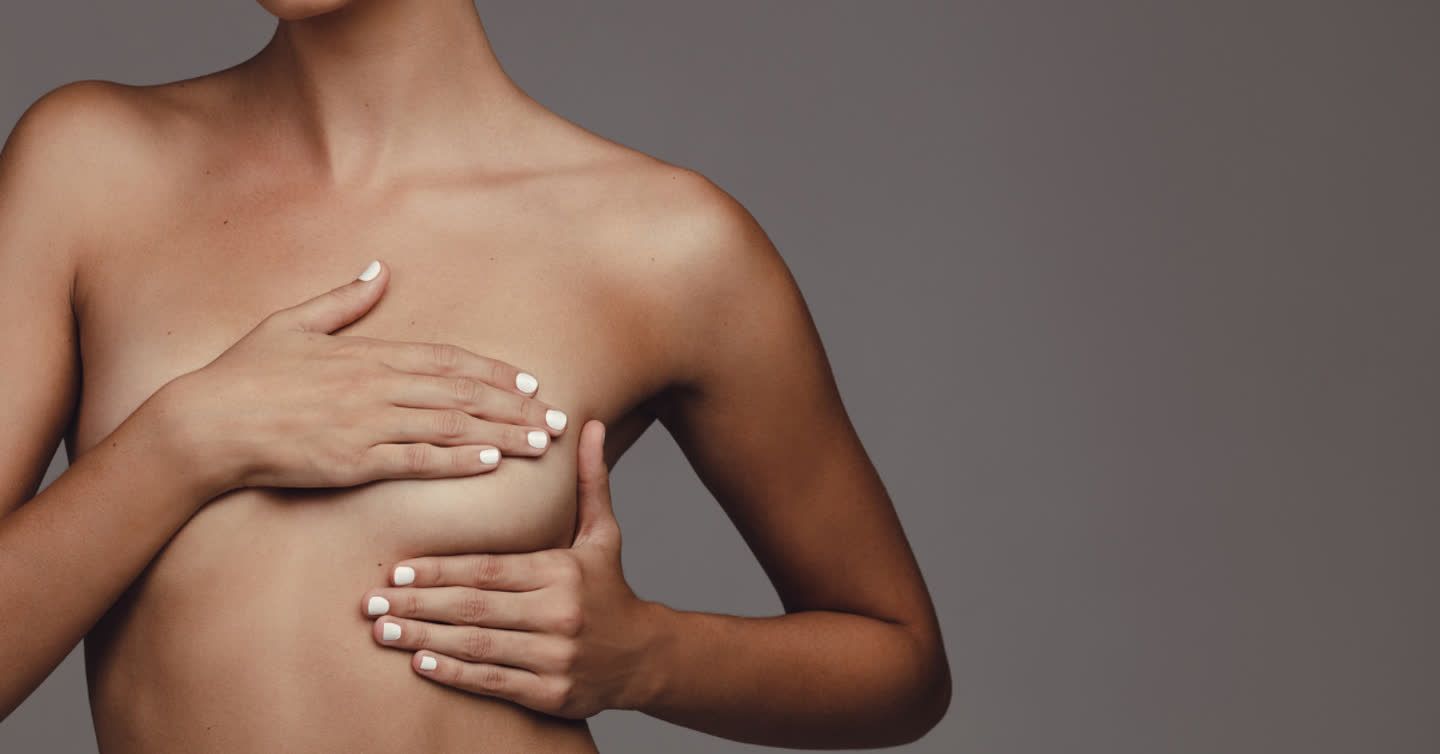 Woman with hands covering her chest against a grey backdrop, illustrating breast lift consultation