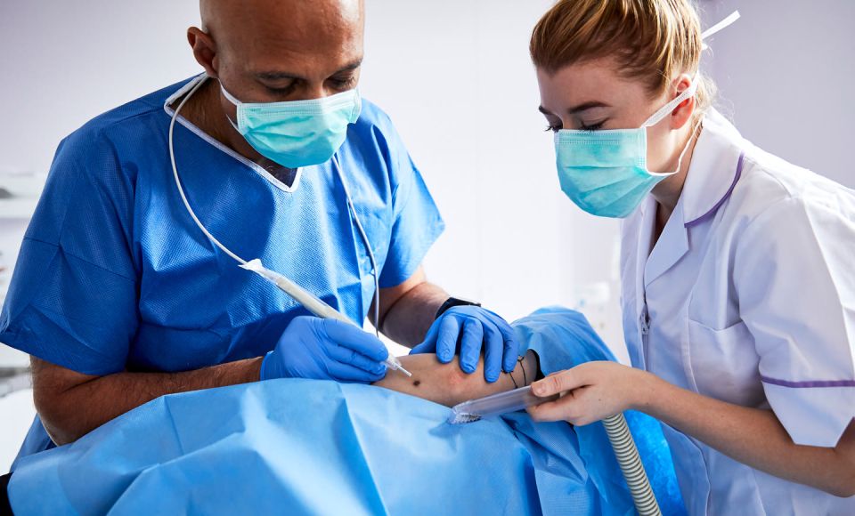 Two masked medical professionals in scrubs performing a minimally invasive body contouring procedure on a patient draped in blue surgical cloth
