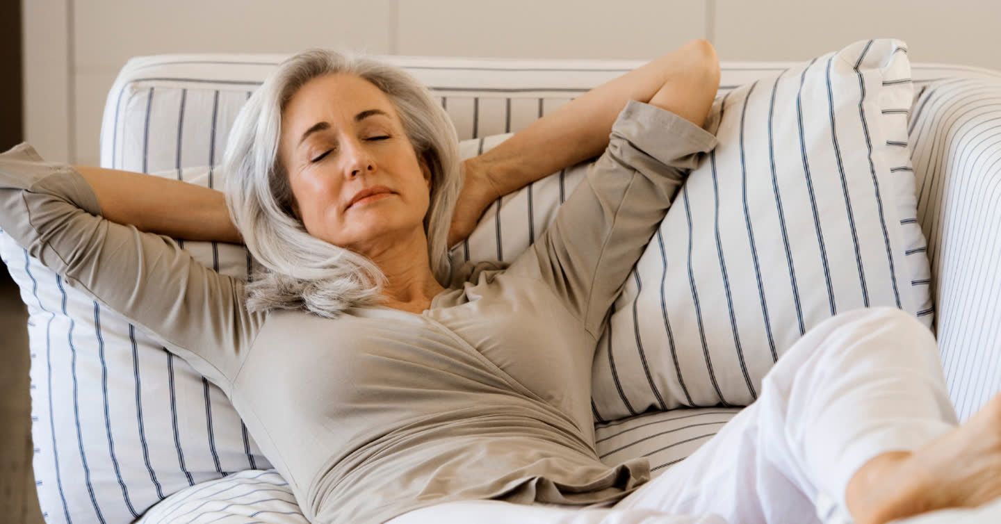 Older woman with grey hair reclining on a striped sofa with eyes closed and arms behind her head