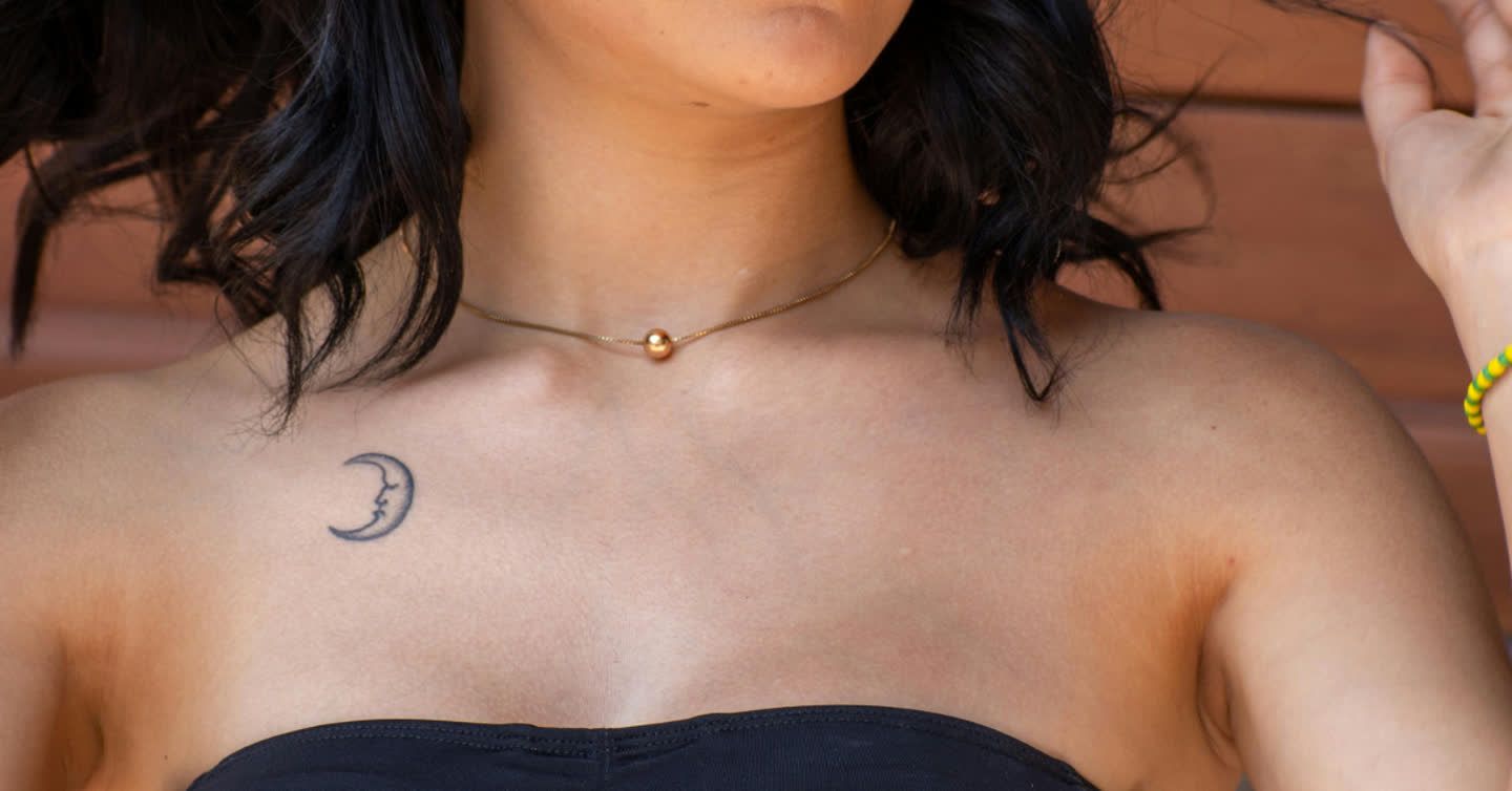 Close-up of a woman's collarbone area showing a small crescent moon tattoo, gold choker necklace, and black strapless top
