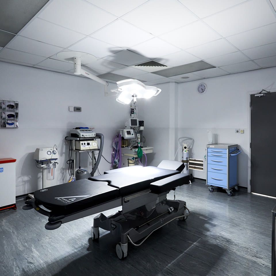 Empty operating theatre with surgical table under bright overhead light, anaesthesia equipment, and blue supply trolley