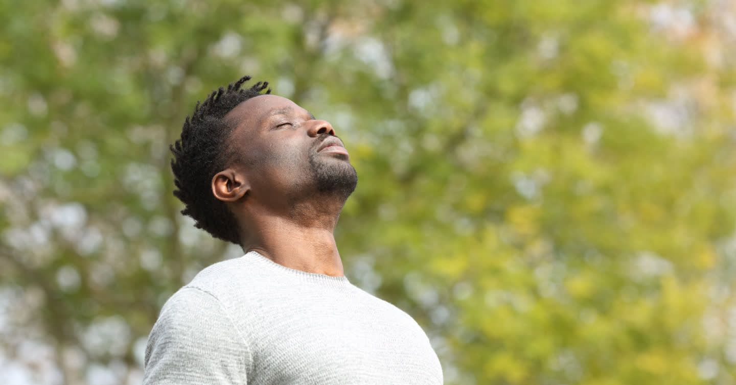 Man with eyes closed tilting his head back and breathing deeply outdoors surrounded by green trees