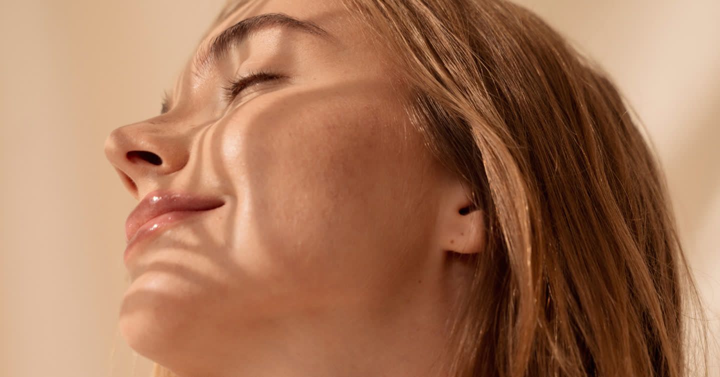 Close-up of a woman with eyes closed tilting her head back, showing smooth glowing facial skin and honey-blonde hair