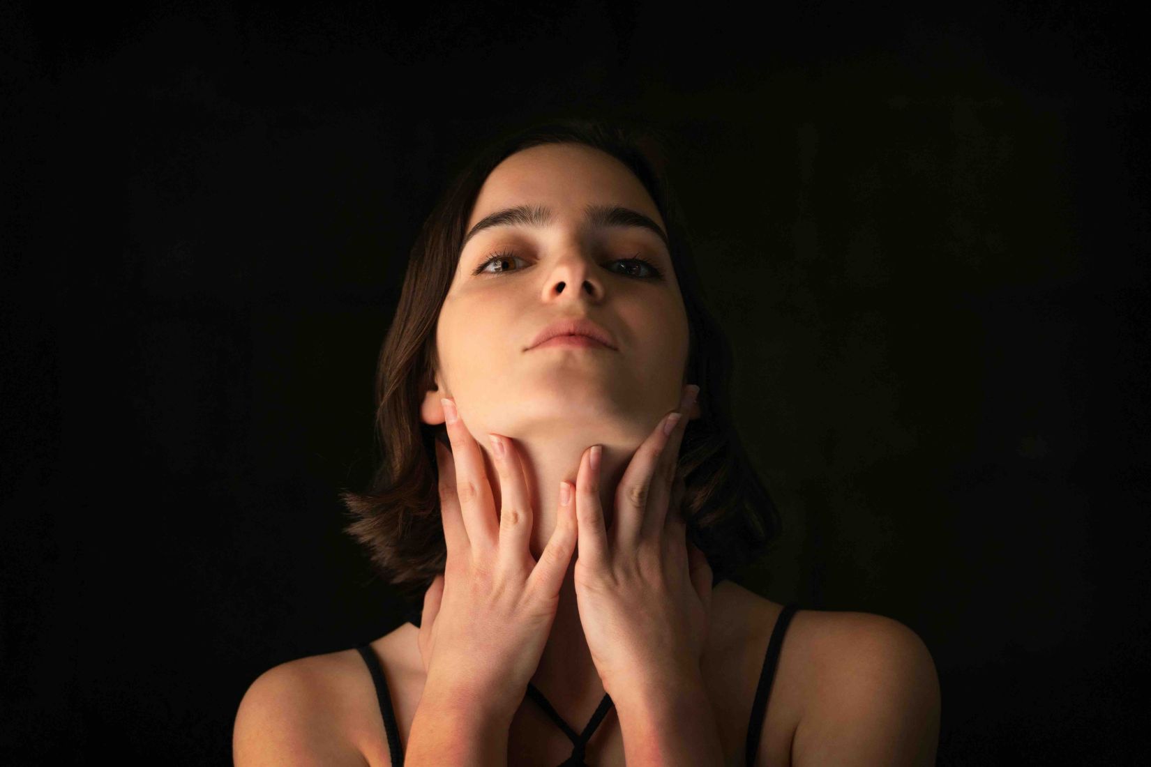 Young woman with both hands gently placed along her jawline and neck against a dark background