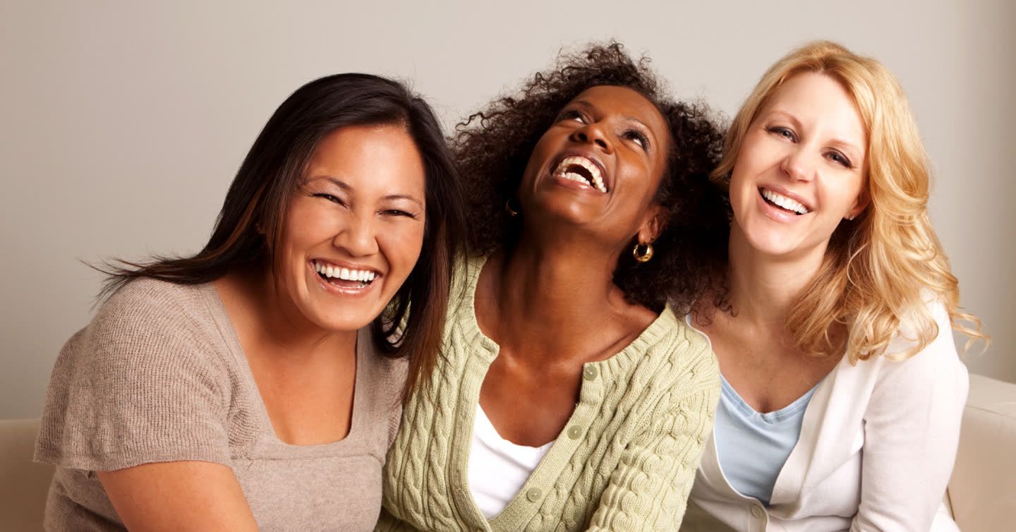 Three women laughing together, leaning into each other with joyful expressions against a neutral background