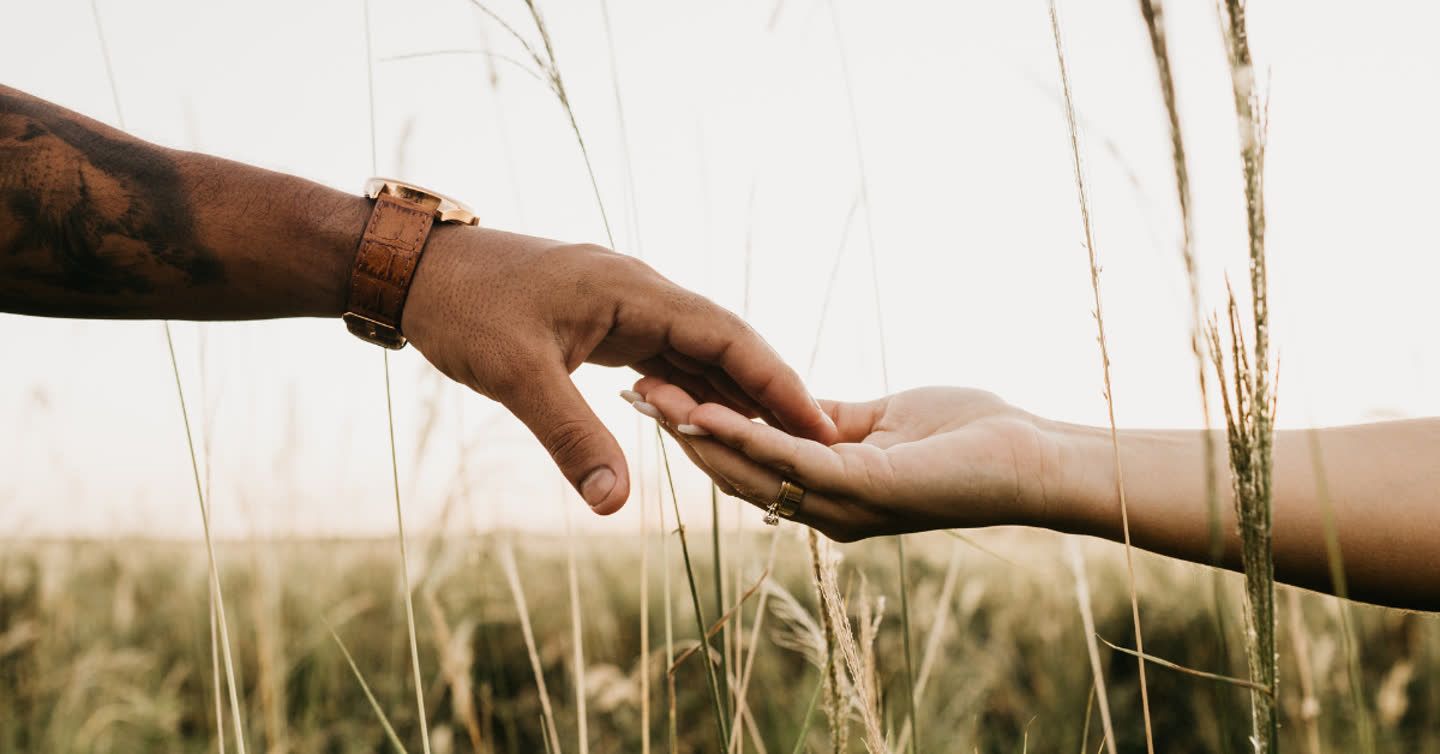 Two hands gently reaching towards each other in a tall grass field, one wearing a leather watch and the other a gold ring